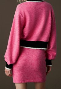 Femme portant un pull en maille rose vif et une jupe assortie, toutes deux avec des poignets et une ceinture à rayures noires et blanches, vue de dos.