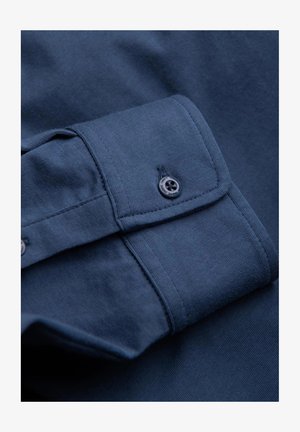 Mörkblå bomullsskjorta med detalj av slät textur och knappmanchetter. Har en enkel svart knapp och förstärkta sömmar.