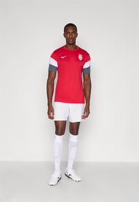Camiseta de fútbol roja con mangas grises y blancas, combinada con pantalones cortos blancos y calcetas blancas hasta la rodilla. Zapatos blancos con tacos negros.