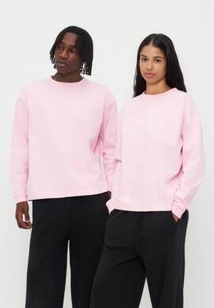 Deux adultes debout côte à côte portant des chemises à manches longues rose clair assorties et des pantalons noirs sur un fond uni.