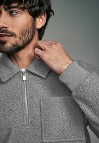 Harmaa fleece-takki, jossa on vetoketjullinen etuosa, muotoiltu kaulus ja rintatasku. Resorikujat sekä huomattava teksturoitu kangas.