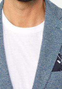 Blå texturerad kavaj med en vit t-shirt under. Kavajen har en mönstrad näsduk i fickan och knäppta slag.