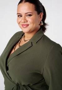 Olijfgroen blazer met een wikkelontwerp, zachte textuur en puntige kraag. Gouden statement oorbellen en een gelaagde ketting voegen detail toe.
