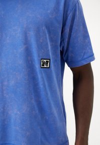 Blue cotton T-shirt with a distressed texture. Features a black and white Nike logo patch on the left chest. Short sleeves.