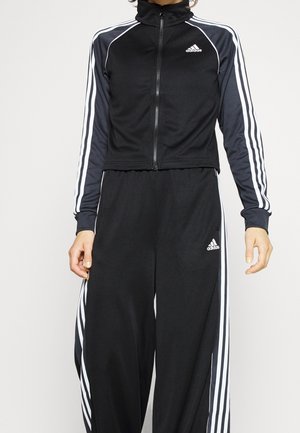 Osoba ubrana w czarny dres marki Adidas z białymi paskami na rękawach i spodniach, stojąca na jednolitym jasnym tle.