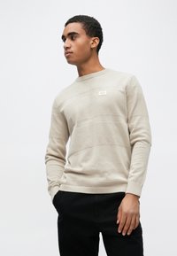 Beež kudumistrikoo tekstuursete horisontaalsete triipudega, ümmarguse kaelusega ja väikese sildiga rinnal. Sobitatud mustade pükstega.