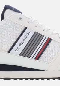 Sneaker bianco con parte superiore tessurizzata, sovrapposizione in tessuto a strisce blu navy, grigio e rosso, con linguetta imbottita e dettaglio del logo.
