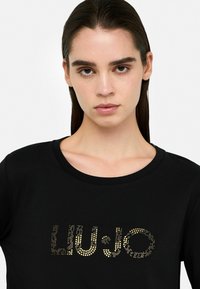 Musta T-särk, millel on ümmargune kaelus ja kuldsete studidega "LIU JO" logo ees, puhta valge tausta taustal.