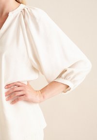 Lys beige, tekstureret top med en afslappet pasform, blødt stof, pufærmer og en samlet halsudskæring, der har et enkelt, minimalistisk design.
