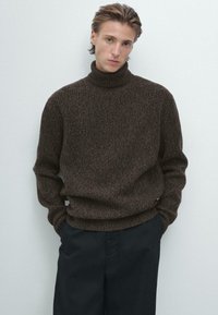 Brązowy sweter z golfem, dziergany, o teksturowanym wykończeniu i luźnym kroju. Nosi się go z ciemnymi spodniami, które mają ściągacze na mankietach i u dołu.