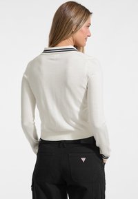 Witte gebreide trui met een getailleerde taille en een zwartgestreepte kraag, gecombineerd met zwarte cargobroek met opgestikte zakken en een driehoekig logo.