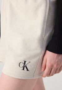Lys grå bomullsshorts med teksturfinish, med en svart "CK" logo nær kanten og en kontrasterende svart ribbestrikket detalj på siden.