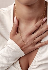 Złoty naszyjnik z wydłużonymi owalnymi ogniwami, spoczywający na szyi. Ręka noszącego delikatnie dotyka naszyjnika. Gładka, błyszcząca tekstura.