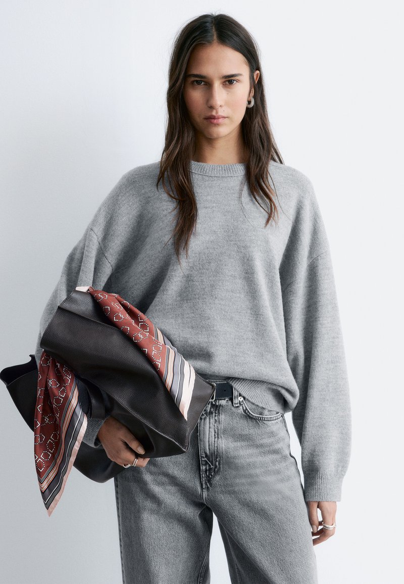 Kvinde med langt mørkt hår iført grå oversized sweater og falmede grå jeans, holder en sort taske med rødt mønstret tørklæde mod en hvid baggrund.