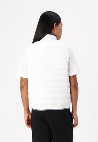 Baltas pūstas liemenė su aukštu apykakle, horizontaliu siuvinėjimu ir raukšlėtu apdailu. Dėvima virš šviesios marškinėlių su trumpomis rankovėmis ir juodų kelnių.