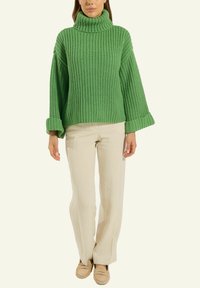 Frau in einem grünen grob gestrickten Rollkragenpullover, beigen weit geschnittenen Hosen und beigen Mokassins, die vor einem einfarbigen hellen Hintergrund steht.