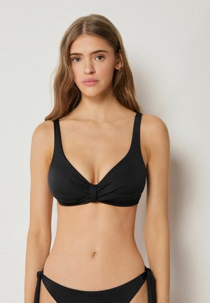 Jeune femme aux longs cheveux portant un bikini noir, haut et bas, sur un fond clair uni, faisant face à l'avant avec une expression neutre.