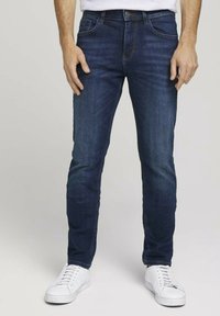 Jeans de mezclilla azul oscuro con corte ajustado, cinco bolsillos y trabillas para cinturón, combinados con zapatillas blancas. Textura suave y diseño clásico.