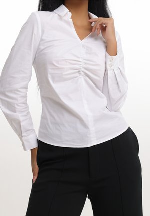 Femme portant un chemisier blanc ajusté à manches longues avec un centre froncé et un pantalon noir, posant avec une main près du col et l’autre dans la poche.
