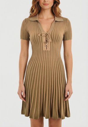 Femme portant une robe courte à manches courtes en maille côtelée beige avec un col et un détail à lacets au niveau du col en V, debout contre un fond uni.