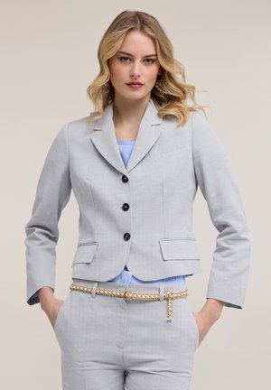 Blonde Frau in hellgrauem Blazer und passender Hose mit einem goldenen Kettengürtel, Hände in den Taschen, neutraler Hintergrund.