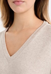 Beżowy sweter z dekoltem w kształcie litery V, wykonany z miękkiego, teksturowanego materiału. Dekolt jest szeroki, a tkanina ma subtelny, melanżowy wzór.