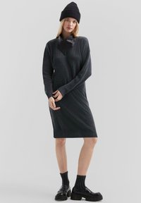 Strickkleid in Dunkelgrau mit gerippter Textur und langen Ärmeln. Verfügt über einen Kragen aus Kunstleder und eine figurbetonte Passform, kombiniert mit schwarzen klobigen Schuhen.