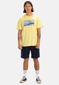 Camiseta de algodón amarilla con un diseño gráfico azul, combinada con pantalones cortos cargo azul marino. El modelo lleva zapatillas negras y calcetines a rayas blancas.