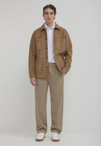 Giacca in suede marrone con due tasche frontali, abbinata a una camicia bianca e pantaloni beige. Il modello indossa sneakers bianche, in piedi davanti a uno sfondo neutro.
