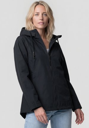 Blonde Frau trägt eine schwarze Kapuzenjacke mit Reißverschluss und hellblaue Jeans, blickt direkt in die Kamera mit neutralem Gesichtsausdruck.