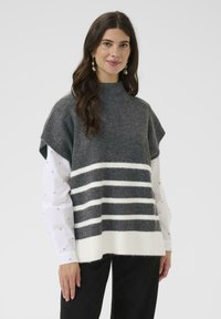 Sieviete ar gariem tumšiem matiem, kas valkā pilienu formas auskari, pelēku bezpiedurkņu džemperi ar baltām svītrām virs baltas garām piedurknēm krekla un melnām biksēm.