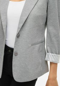 Persona che indossa un blazer grigio con polsini a righe su una camicia bianca e pantaloni neri, che mostra dettagli di bottoni e tasche.