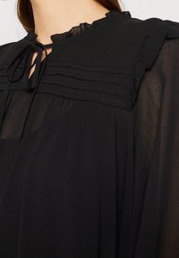 Blouse noire avec un col froncé et des détails plissés, présentant une ouverture en forme de trou de serrure au niveau du cou et une texture de tissu transparent et fluide.