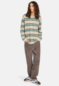 Camicia beige a maniche lunghe con righe orizzontali turchesi e verde scuro, abbinata a pantaloni marroni larghi e sneakers multicolori.