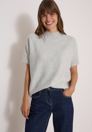 Femme souriante portant un pull en maille gris clair à manches courtes et un jean bleu foncé, une main dans sa poche, sur un fond uni.