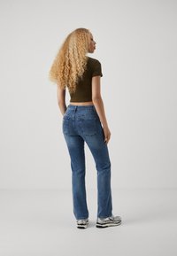 Blue denim flared jeans with a light wash, five pockets, and a brown tag at the waistband, worn with a fitted brown top and silver sneakers.
