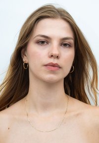 Goldene Creolen und eine filigrane goldene Kettenhalskette schmücken ein Model mit langen, welligen Haaren, vor einem schlichten weißen Hintergrund.