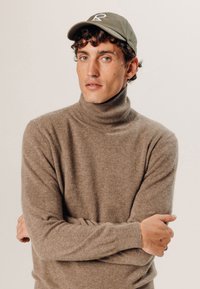 Bruine coltrui gemaakt van zacht materiaal, gecombineerd met een groene pet met een wit logo. De pasvorm is relaxed met lange mouwen.
