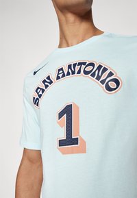Svetlomodré bavlnené tričko s nápisom "SAN ANTONIO" na prednej strane v tučnom námorníckom a broskyňovom písmene, s veľkou grafikou "1" v námorných bodkách. Logo Nike je prítomné.