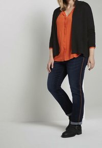 Černý cardigan s rukávy po lokty přes oranžovou košili s knoflíky, ve spojení s tmavými džíny se oranžovým prošíváním po stranách a černými kotníkovými botami.
