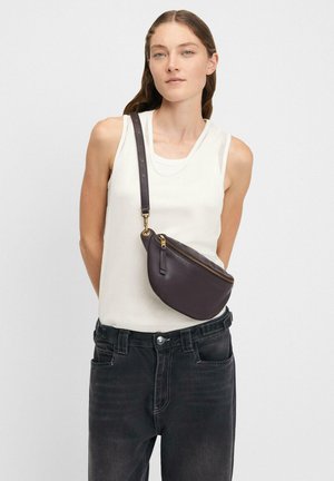 Junge Frau mit ärmellosem weißen Top, schwarzen Jeans und einer dunklen Leder-Crossbody-Tasche mit goldener Reißverschluss.