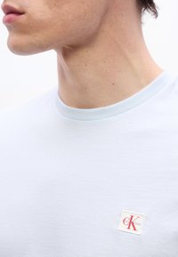 Κοντινό πλάνο του λαιμού και του σαγονιού ενός ατόμου που φοράει ένα γαλάζιο μπλουζάκι Calvin Klein με ένα μικρό κόκκινο λογότυπο CK στο στήθος.