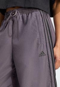 Heledad lilla spordipüksid mustade külgtriipudega. Elastne vöökoht koos nööriga, millel on must tikandiga Adidas logo.
