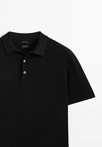 Polo-shirt noir avec un motif de tricot texturé, manches courtes, un col classique et une patte de boutonnage à trois boutons. Étiquette visible à l'intérieur du col.