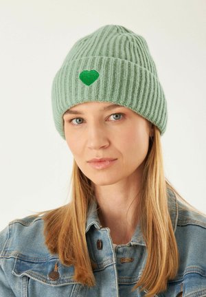 Bonnet tricoté à côtes de couleur vert clair, orné d'une broderie en forme de cœur vert à l'avant. Porté avec une veste en jean, mettant en valeur son design décontracté.