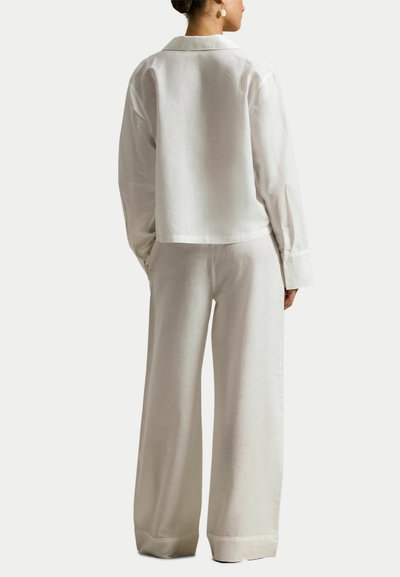 Vrouw staande met de rug naar de kijker, draagt een losse witte blouse met lange mouwen en wijde broek, tegen een effen achtergrond.