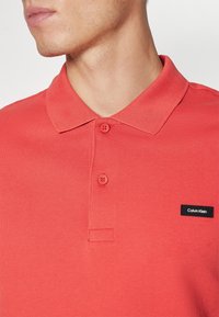Homme portant un polo rouge corail Calvin Klein avec deux boutons fermés et une étiquette de marque visible sur la poitrine.