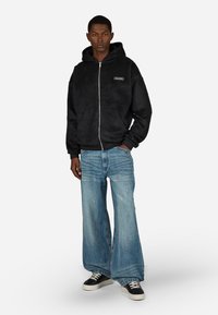 Črna flis jakna z zadrgo in sprednjimi žepi, oblečena z svetlo modrimi širokimi hlačami in črnimi supergami z belimi podplati.