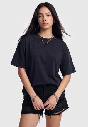 Ung kvinde med langt mørkt hår bærer en sort oversized t-shirt, slidte sorte shorts og sølvkæde smykker med pigge og vedhæng.