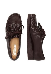 Donkerbruine leren instappers met kwastjes en veters, crepe zool, één schoen van bovenaf met het "Clarks ORIGINALS" binnenzool zichtbaar, andere schoen van opzij.